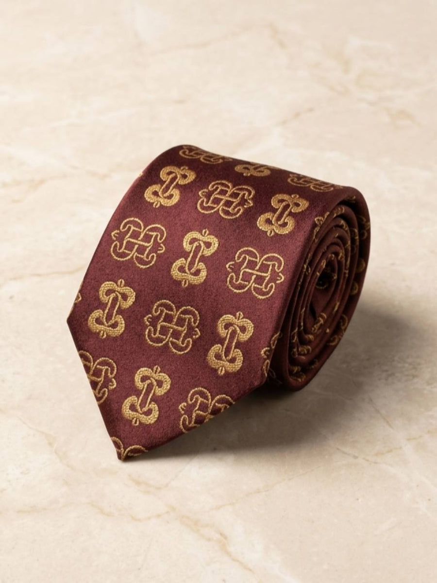 ODEHYE Burgundy Heritage Tie rolled on warm stone, showing aligned gold Adinkra symbols